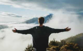 Back view of anonymous male traveler in casual clothes standing on edge of cliff and admiring breathtaking scenery of clouds and mountain top