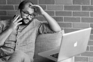 Worried man using a smartphone beside a laptop, illustrating online dating scams and digital fraud